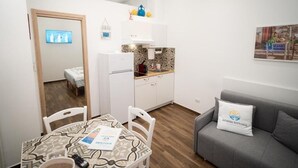 A compact living space with a dining area, kitchenette, and a grey sofa with a pillow. - Nonna Vittoria Apartments (Mola di Bari)