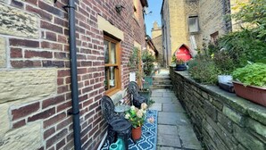 A narrow alleyway with a brick building on one side and a stone wall on the other. There is a small table with chairs and a potted plant in the center. - Sleeps 4 - Old Farmhouse - Near Pendle Hill (Skipton)