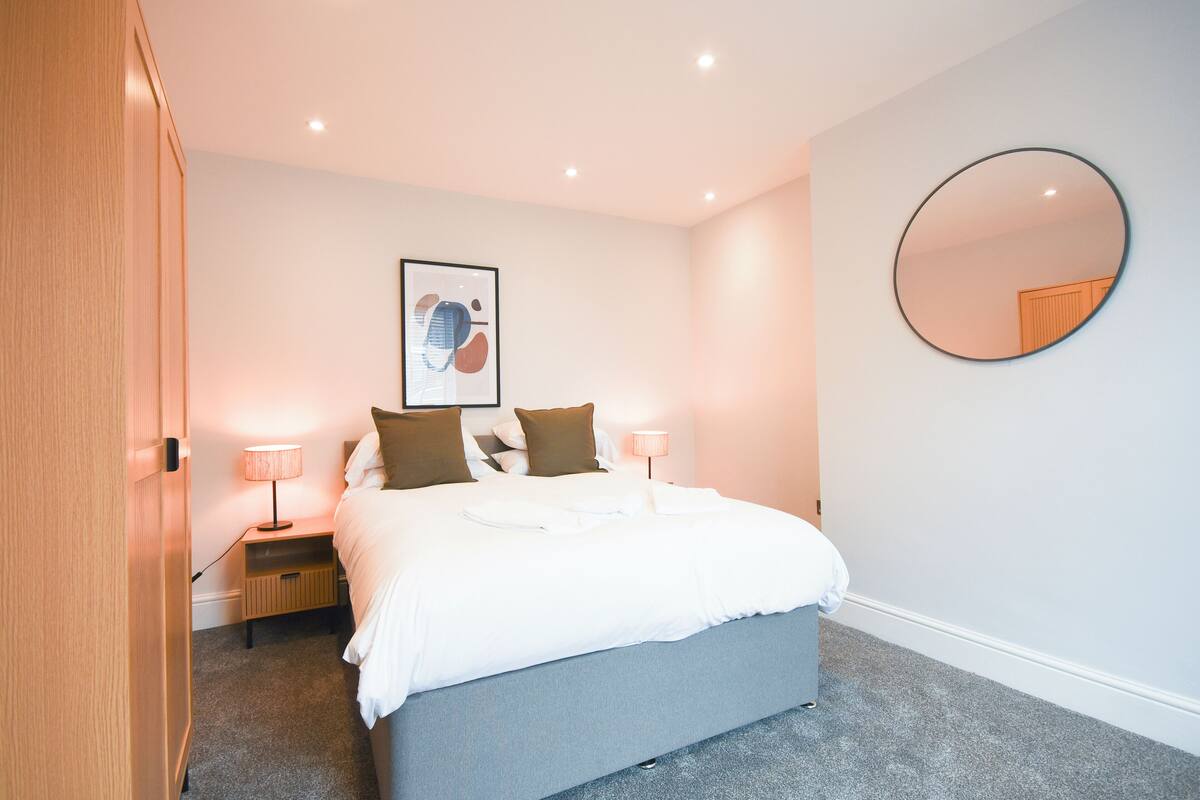 Un lit bien fait, avec du linge de lit blanc, un miroir rond accroché au mur et une œuvre d’art encadrée au-dessus du lit.