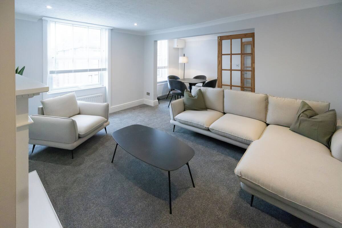 Un salon moderne avec un canapé blanc, un fauteuil assorti et une table basse foncée. On aperçoit une table à manger avec des chaises au fond.