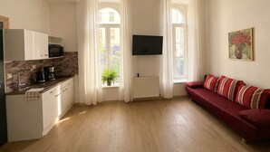 A modern living room with a red sofa, a flat-screen TV mounted on the wall, and a kitchen area with a microwave and coffee maker. - Cozy Apartment for 3 Near Lugner City (Vienna)