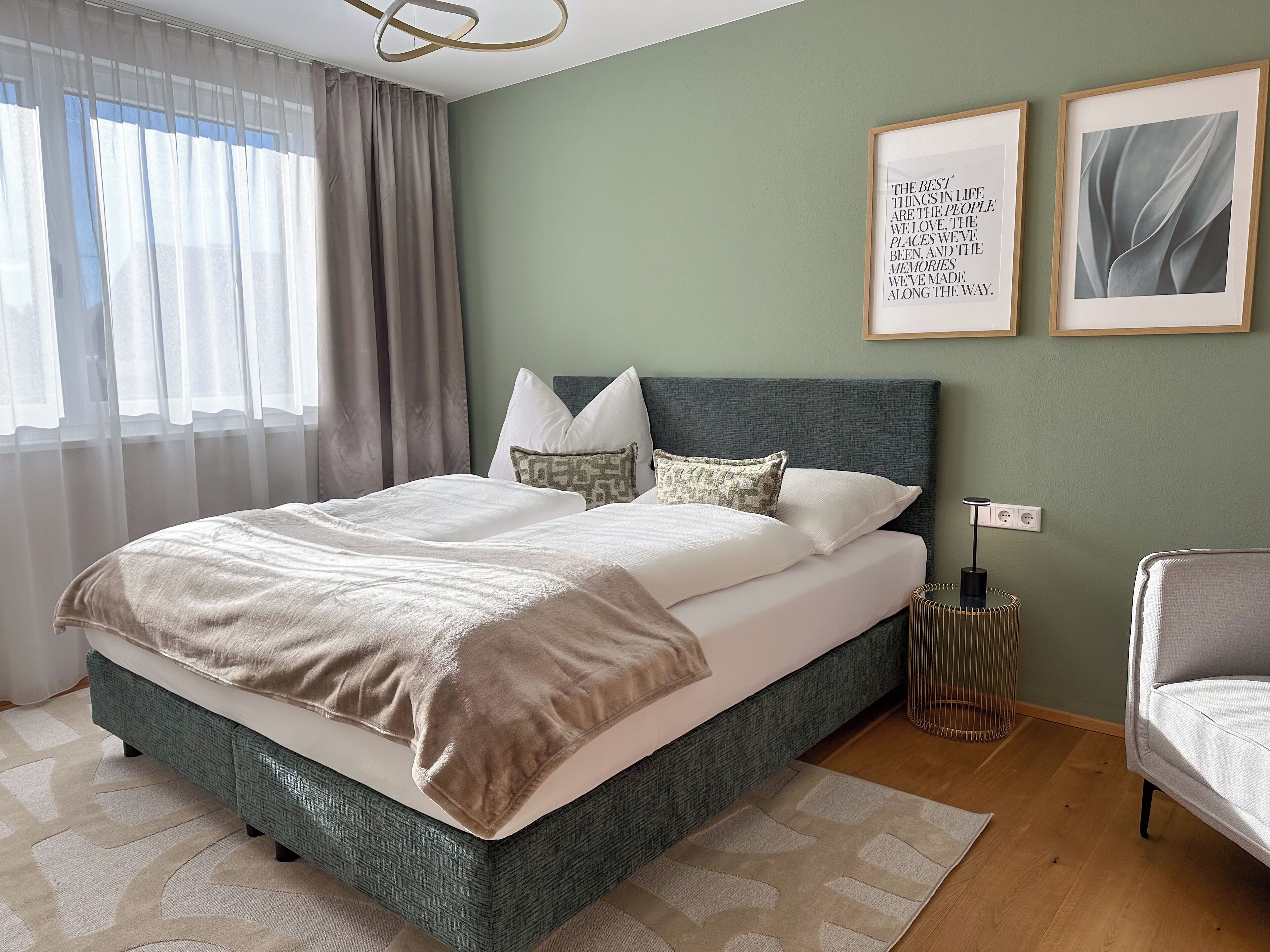 A neatly made bed with a green headboard, a bedside table with a lamp, and two framed quotes on the wall.
