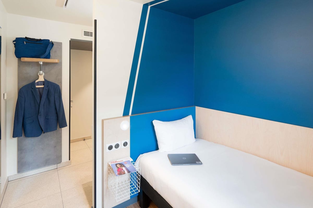 Habitació d'hotel moderna amb una paret d'accent blau, un llit ben fet amb un ordinador portàtil, una estant per a abrics penjants i una petita taula de nit amb una revista.