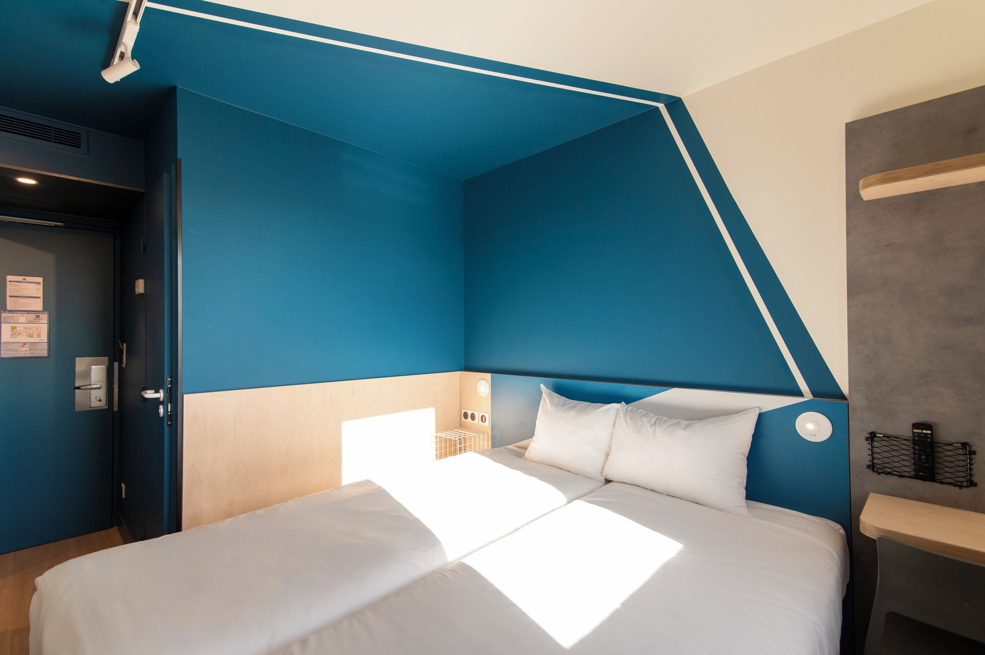 A modern hotel room with a bed, a bedside table, a nightstand, and a built-in shelf.