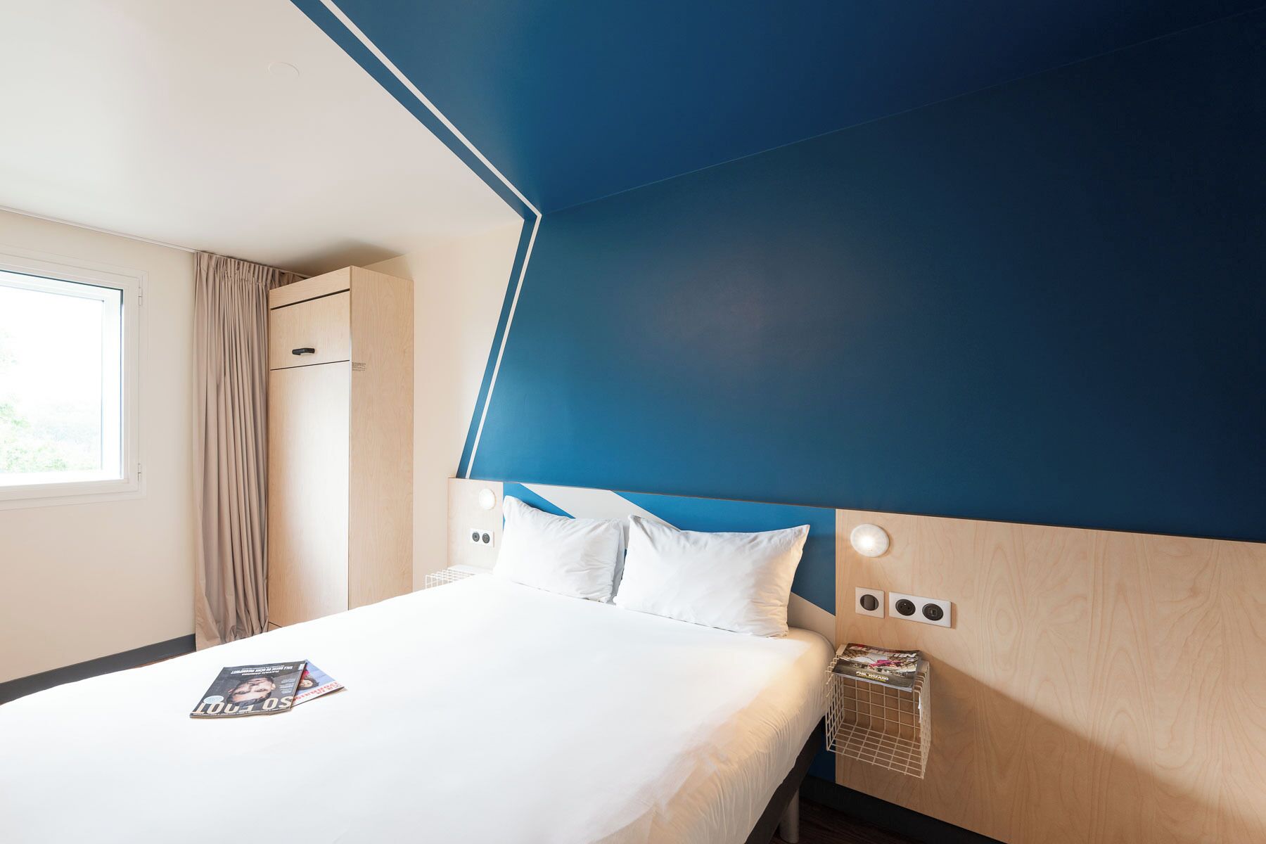 Une chambre d’hôtel avec un lit, une fenêtre avec des rideaux, une tête de lit en bois et une table de chevet sur laquelle se trouve un magazine.