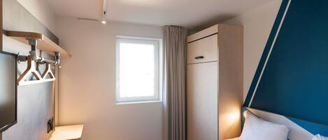 A hotel room with a bed, a window, a wardrobe, and a bedside table.
