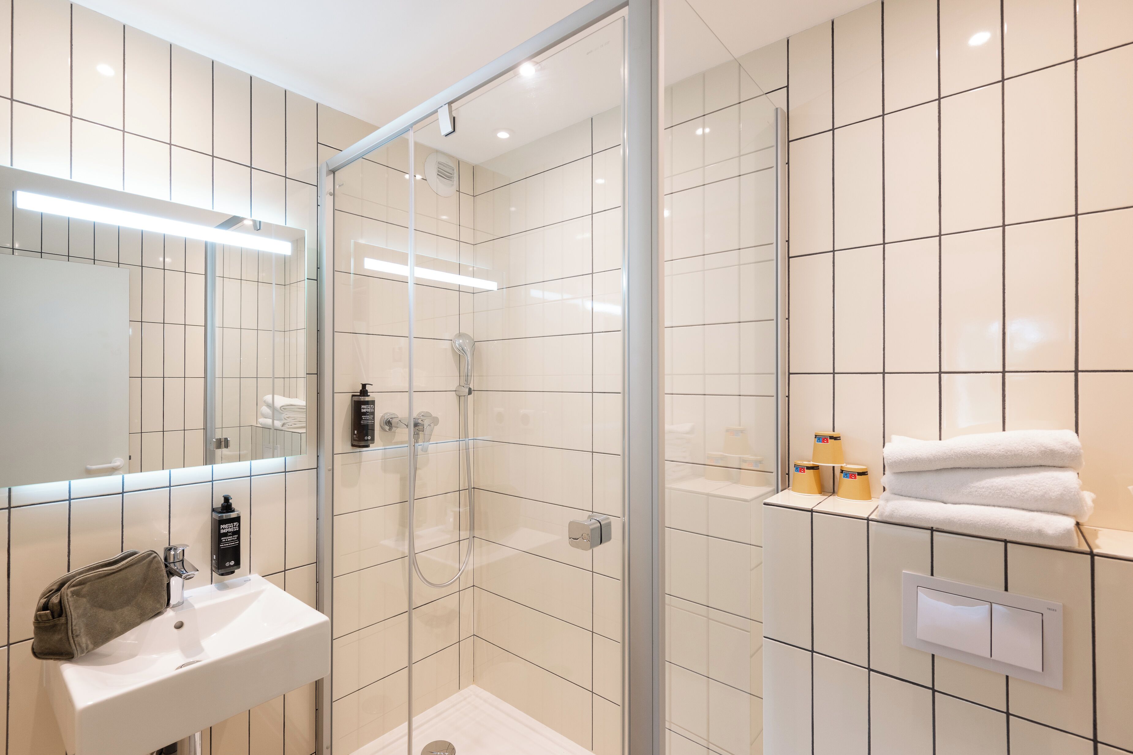 Un baño moderno con ducha de vidrio, lavamanos blanco con artículos de aseo y toallero con toallas dobladas.