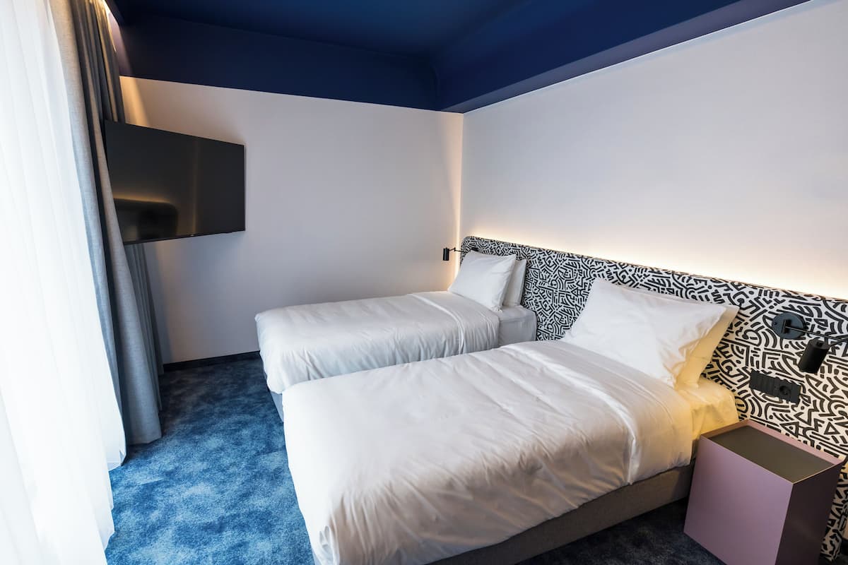 Habitación de hotel con dos camas, un cabecero estampado, un televisor de pantalla plana montado en la pared y una mesita de noche rosa.