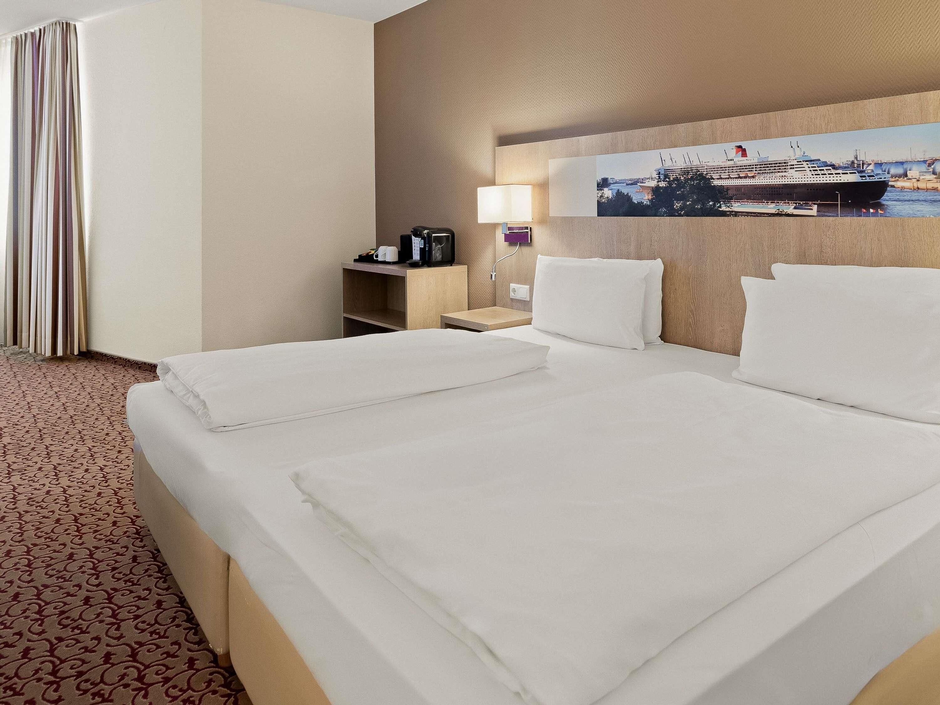 A hotel room with a large bed, a desk with a lamp, a painting on the wall, and a patterned carpet.
