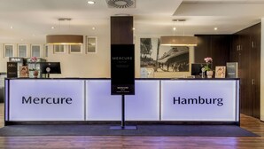 Una mesa de recepción con letreros que indican 'Mercure' y 'Hamburgo', un cartel vertical y una foto enmarcada de un crucero.