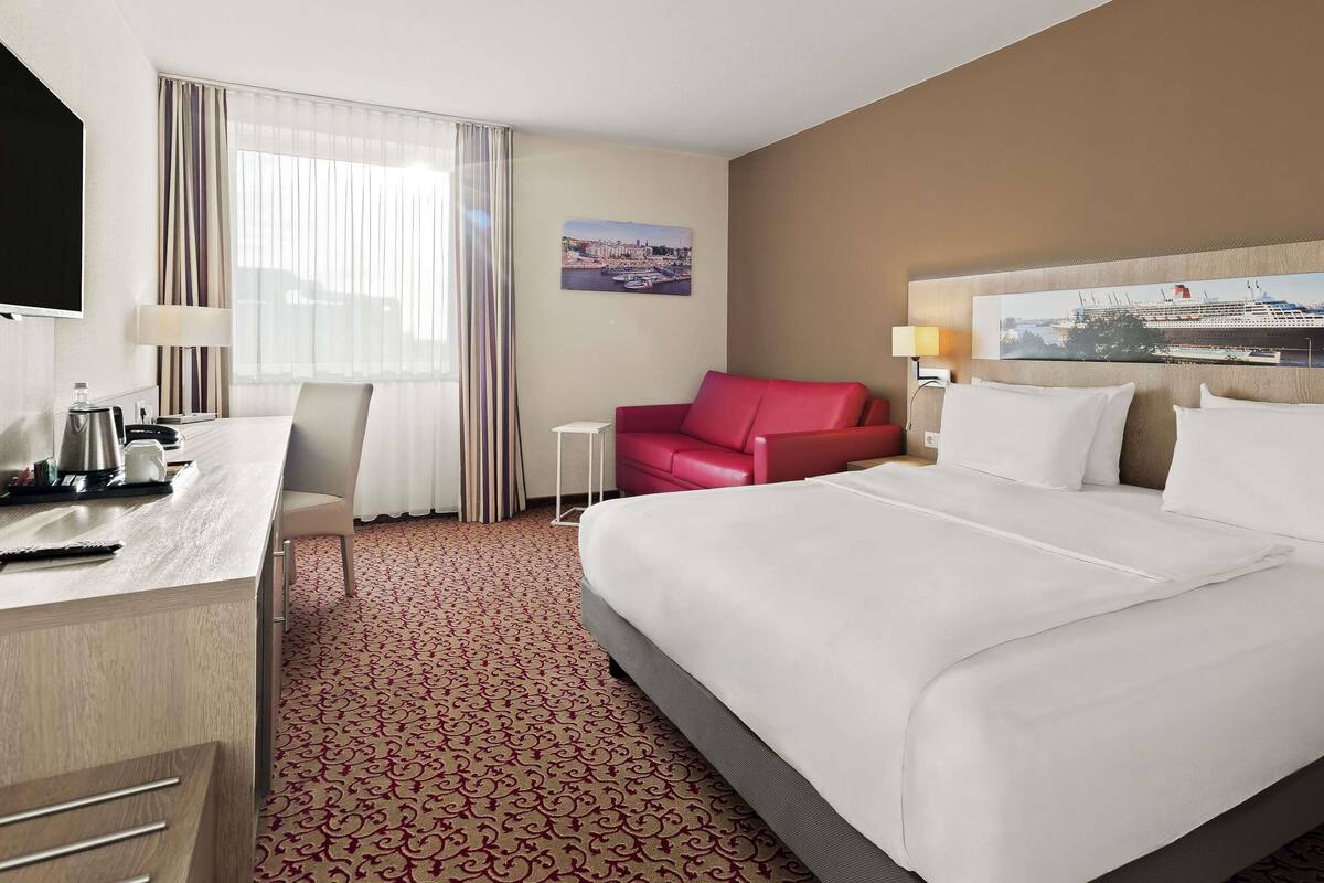 A hotel room with a bed, a desk with a lamp, a chair, a red sofa, and a painting on the wall.