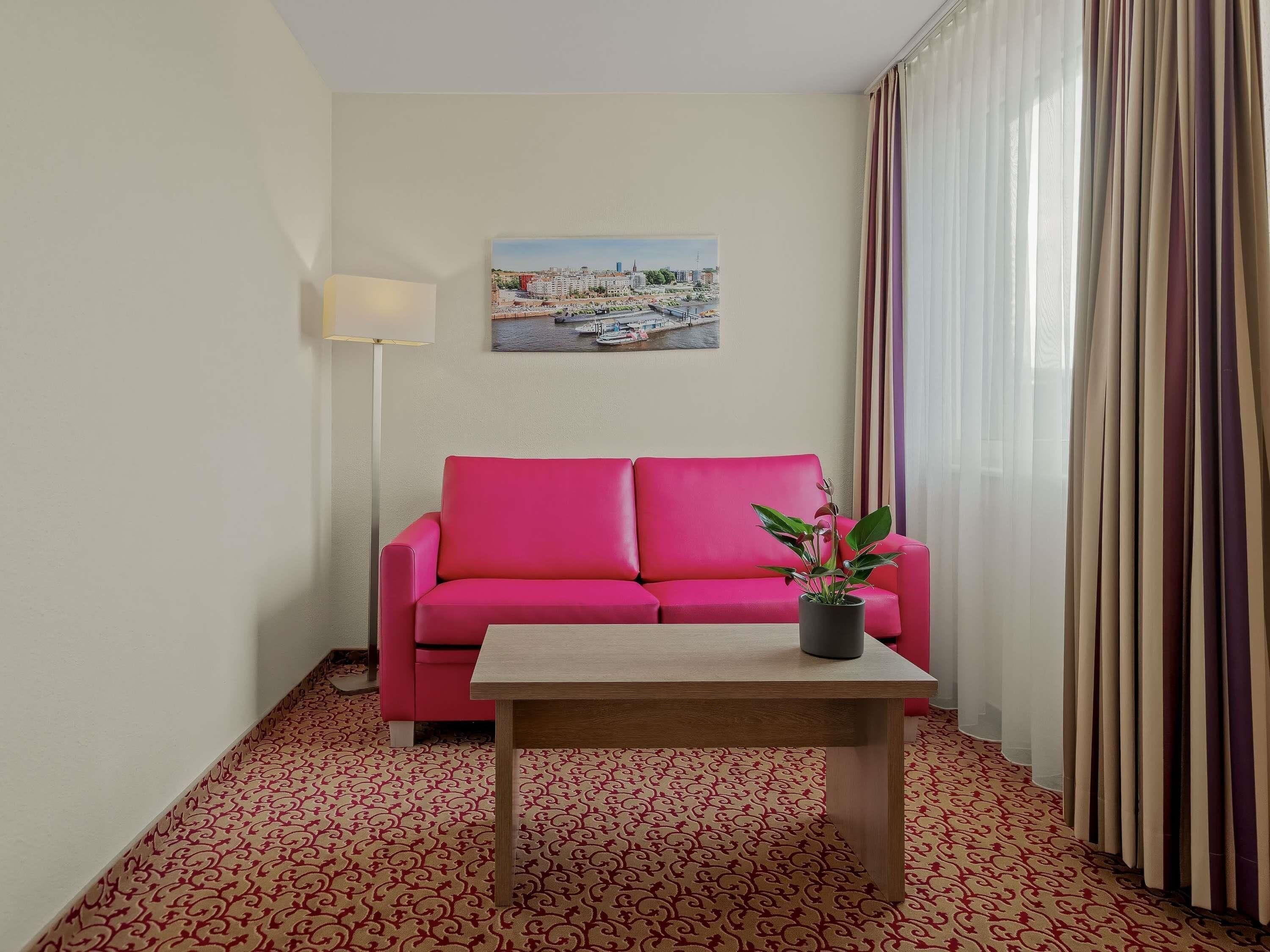 A room with a pink sofa, a small table, a lamp, a painting on the wall, and a patterned carpet.