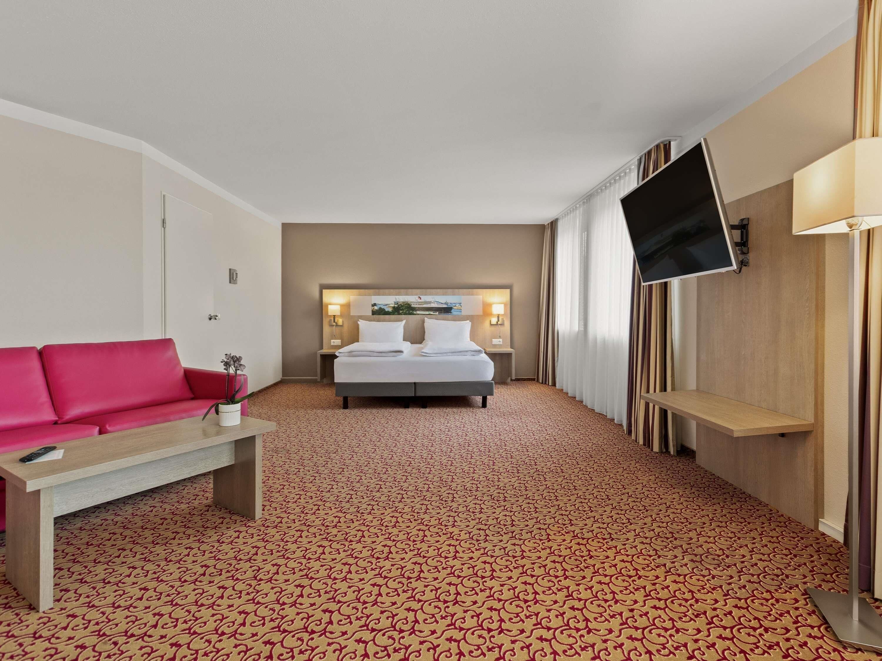 Ein Hotelzimmer mit Bett, Fernseher, rotem Sofa, kleinem Tisch und einem gemusterten Teppich.