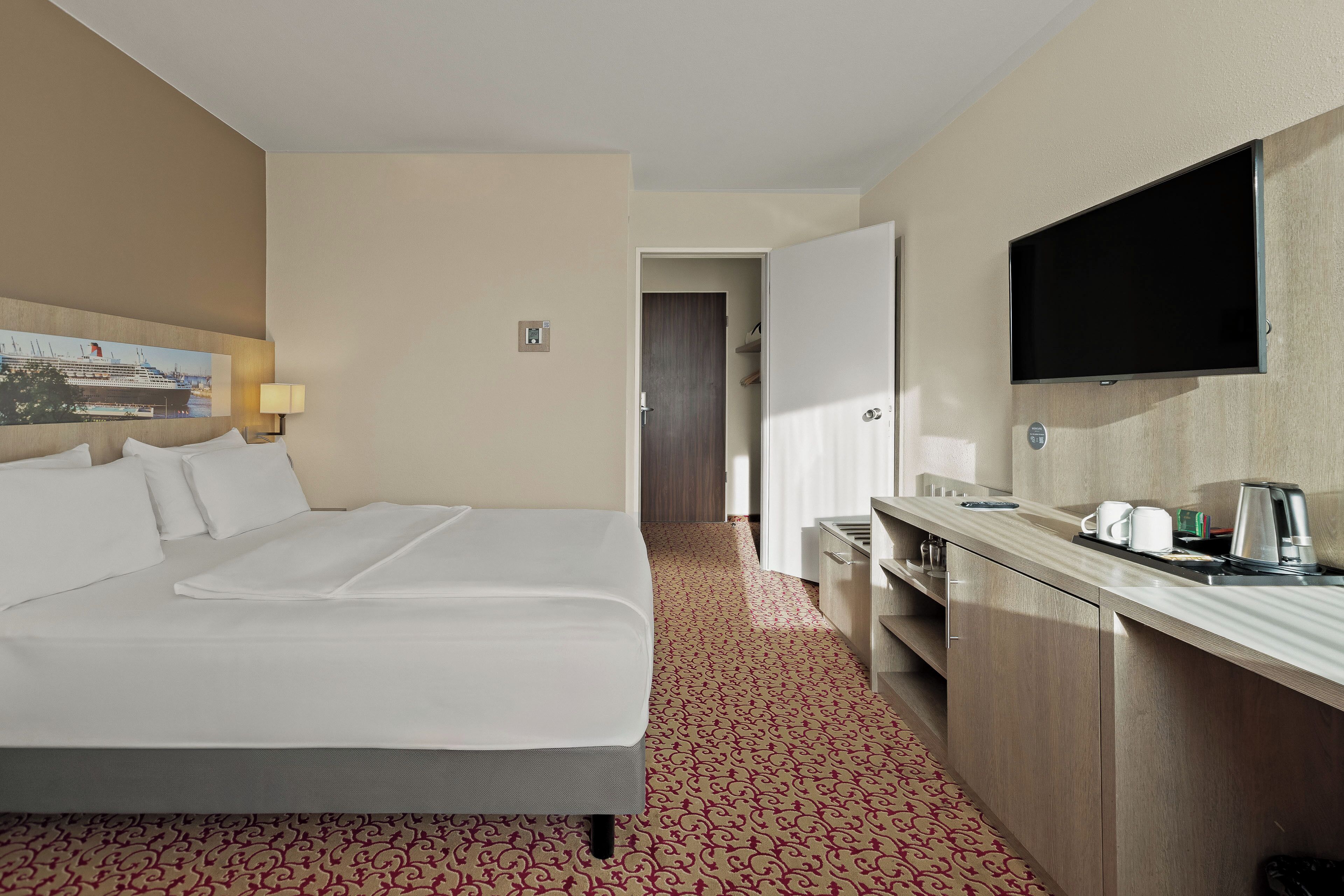 A hotel room with a bed, a TV, a desk with a coffee maker, and a patterned carpet.