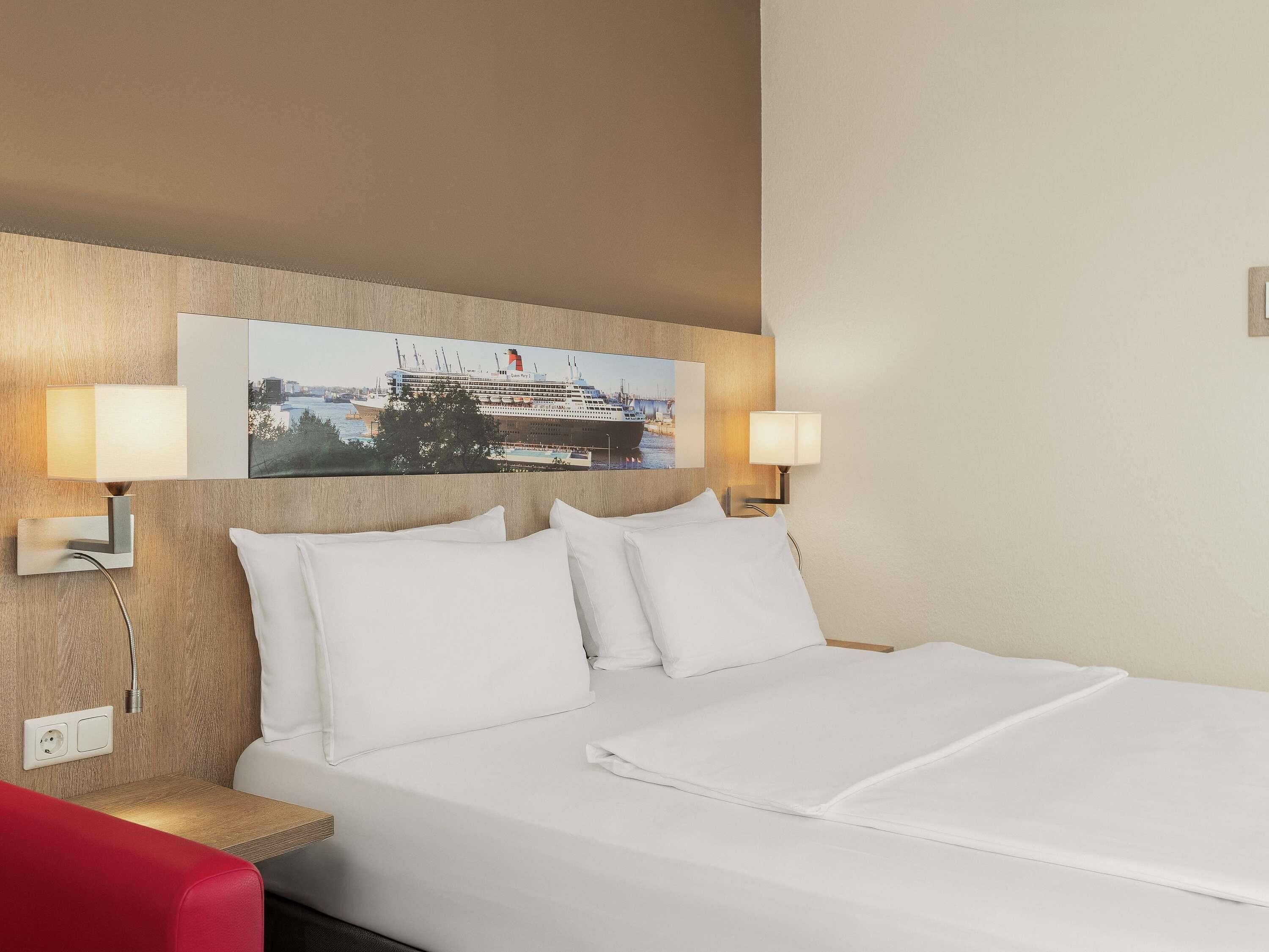 A hotel room with a bed, two wall-mounted lamps, a painting of a cruise ship, and a red chair.