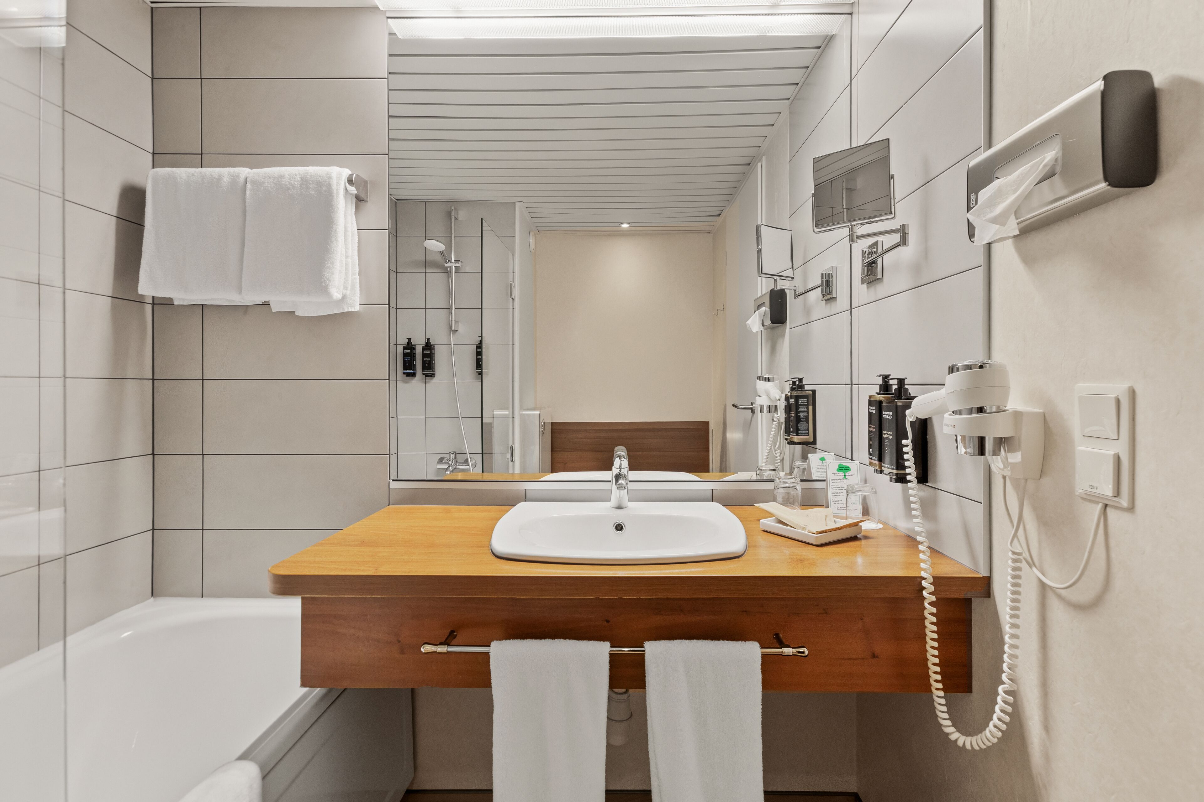 Une salle de bain moderne avec un meuble-lavabo en bois, un lavabo blanc, une baignoire, un miroir, un porte-serviettes et un téléphone.