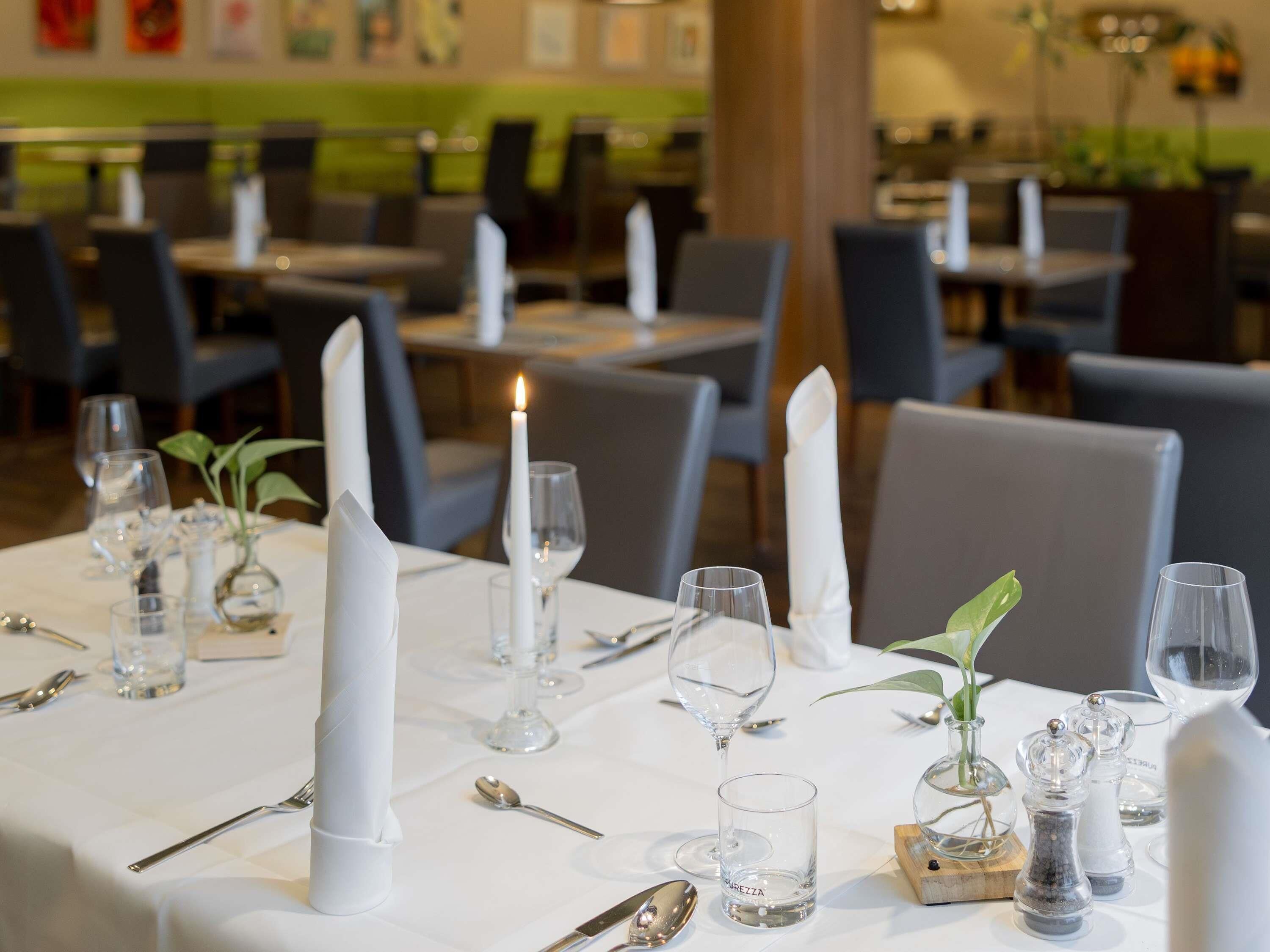 Ambiente de restaurante com mesas preparadas para refeições, com toalhas brancas, velas e copos.