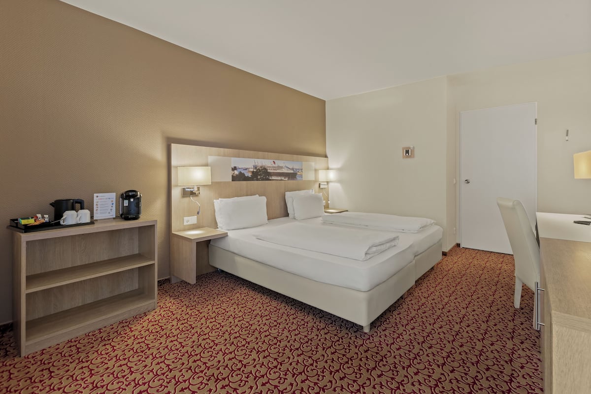A hotel room with a bed, bedside tables, a desk, a chair, a shelf with items, and a patterned carpet.