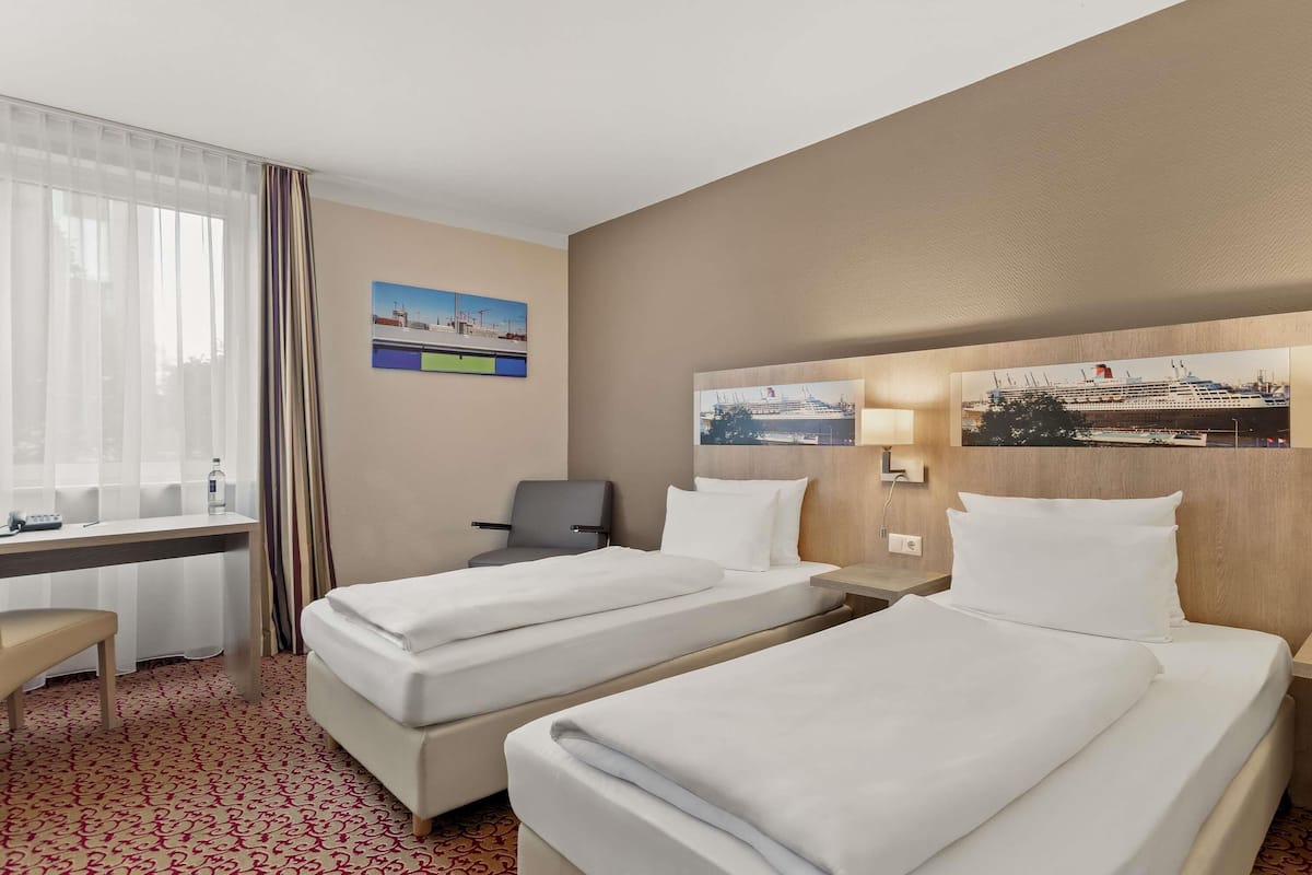 A hotel room with two beds, a desk, a chair, and a window with curtains.