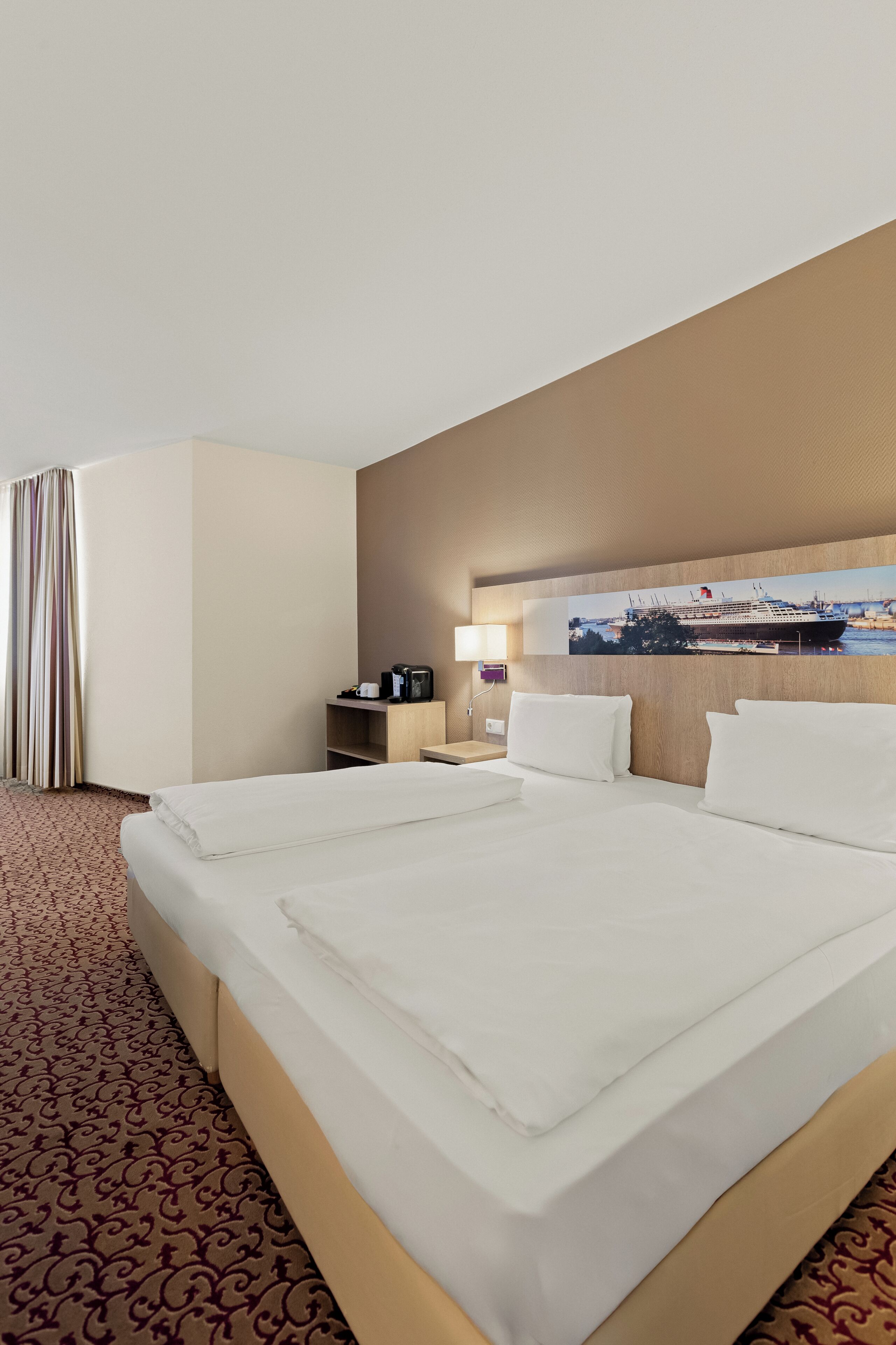 Habitación de hotel con una cama grande, una mesita de noche, una lámpara y un cuadro colgado en la pared con un crucero.