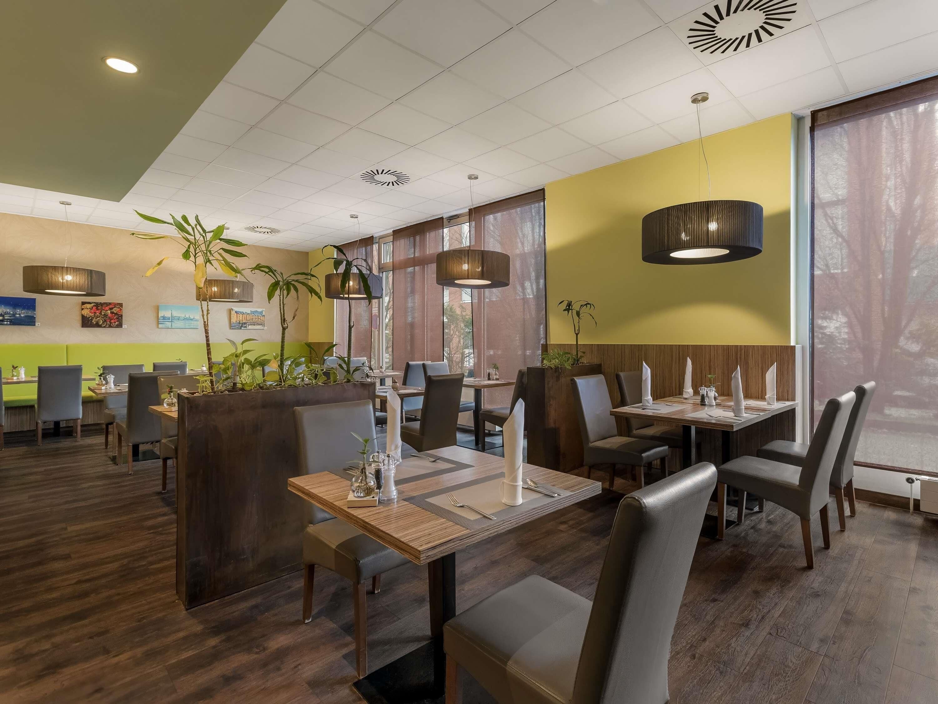 A modern restaurant interior with wooden tables and chairs, potted plants, and pendant lighting.
