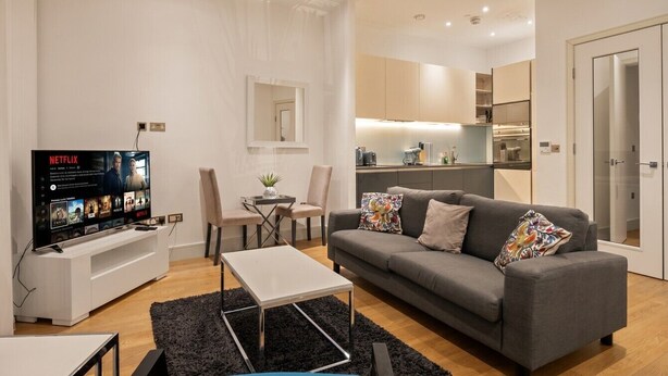 Ein modernes Wohnzimmer mit grauem Sofa, Couchtisch, Fernseher auf weißem Standfuß und einer kleinen Essecke mit Tisch und Stühlen.