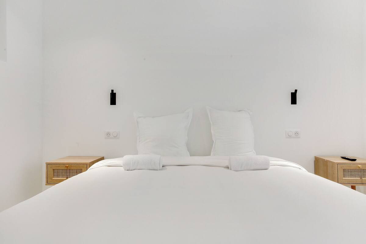 Quarto de hotel minimalista com uma cama grande, duas mesas de cabeceira e uma luminária simples fixada na parede.
