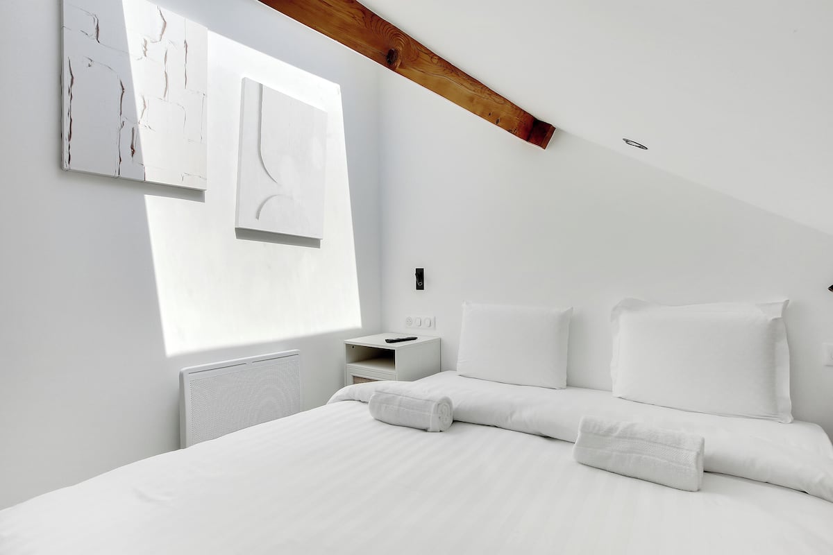 Quarto moderno com cama grande, roupa de cama branca, mesa de cabeceira e janela com obra de arte.