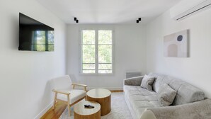 Ein modernes Wohnzimmer mit einem grauen Sofa, einem weißen Sessel und zwei Holztischen. Ein Flachbildfernseher ist an der Wand befestigt, und ein großes Fenster bietet Blick auf Grünflächen.