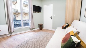 A living room with a sofa, a television mounted on the wall, a window with a view of a building, and a door. - Appartements rue de Fécamp (Paris)