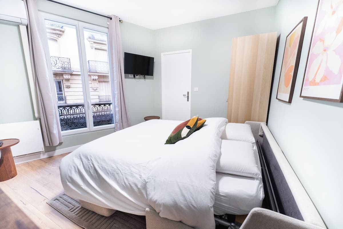 Un dormitorio con cama, televisión montada en la pared, puerta y ventana con cortinas.
