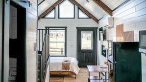 A bedroom with a loft bed, a small table, and a door leading to a balcony. - Natural Nook | Tiny Home #2 in Amish Country! (Shipshewana)