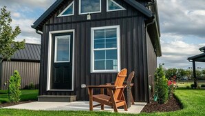A modern two-story house with a dark exterior, large windows, and a front porch with a wooden chair. - Natural Nook | Tiny Home #2 in Amish Country! (Shipshewana)