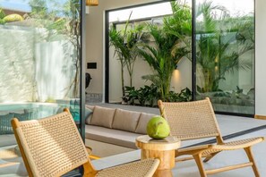 A modern indoor relaxation area with a glass wall, a sofa, and two wicker chairs. - Villa Bayu by GORO (Bali)