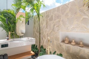 A modern bathroom with a marble sink, a large mirror, a bathtub, and a stone wall. - Villa Bayu by GORO (Bali)