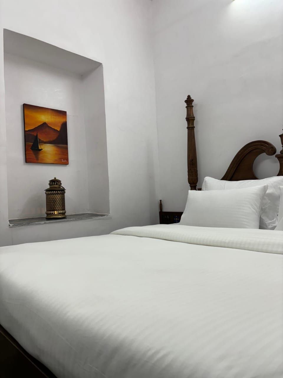 A bedroom with a single bed, a wooden headboard, a nightstand, a wall-mounted shelf with a decorative item, and a painting of a sunset over water.
