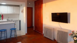 Una cocina funcional con cubierta blanca, dos taburetes azules, una puerta de madera, un televisor montado en la pared y un radiador.