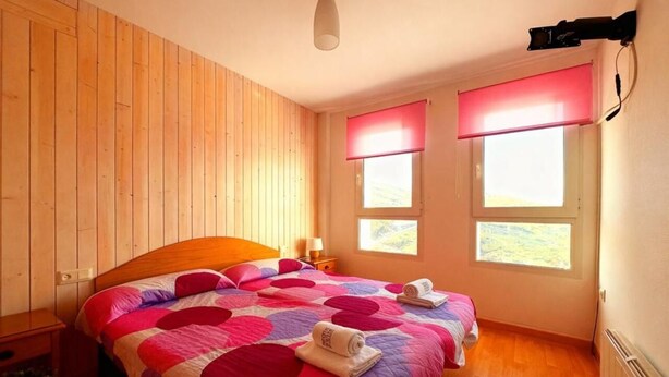 Un dormitorio con pared de paneles de madera, una cama con colcha rosa y blanca, dos ventanas con persianas rojas y un ventilador de techo.