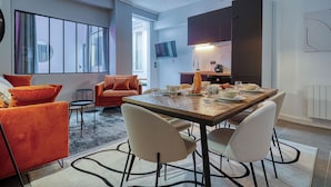 A modern living room with a large window, orange sofas, a dining table set for four, and a kitchen area with cabinets and a TV. - Appartements rue des Jeuneurs (Paris)