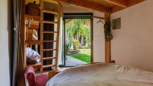 Un dormitorio con cama, una silla de madera, un estante con bolsas y vista a un jardín desde una puerta abierta.