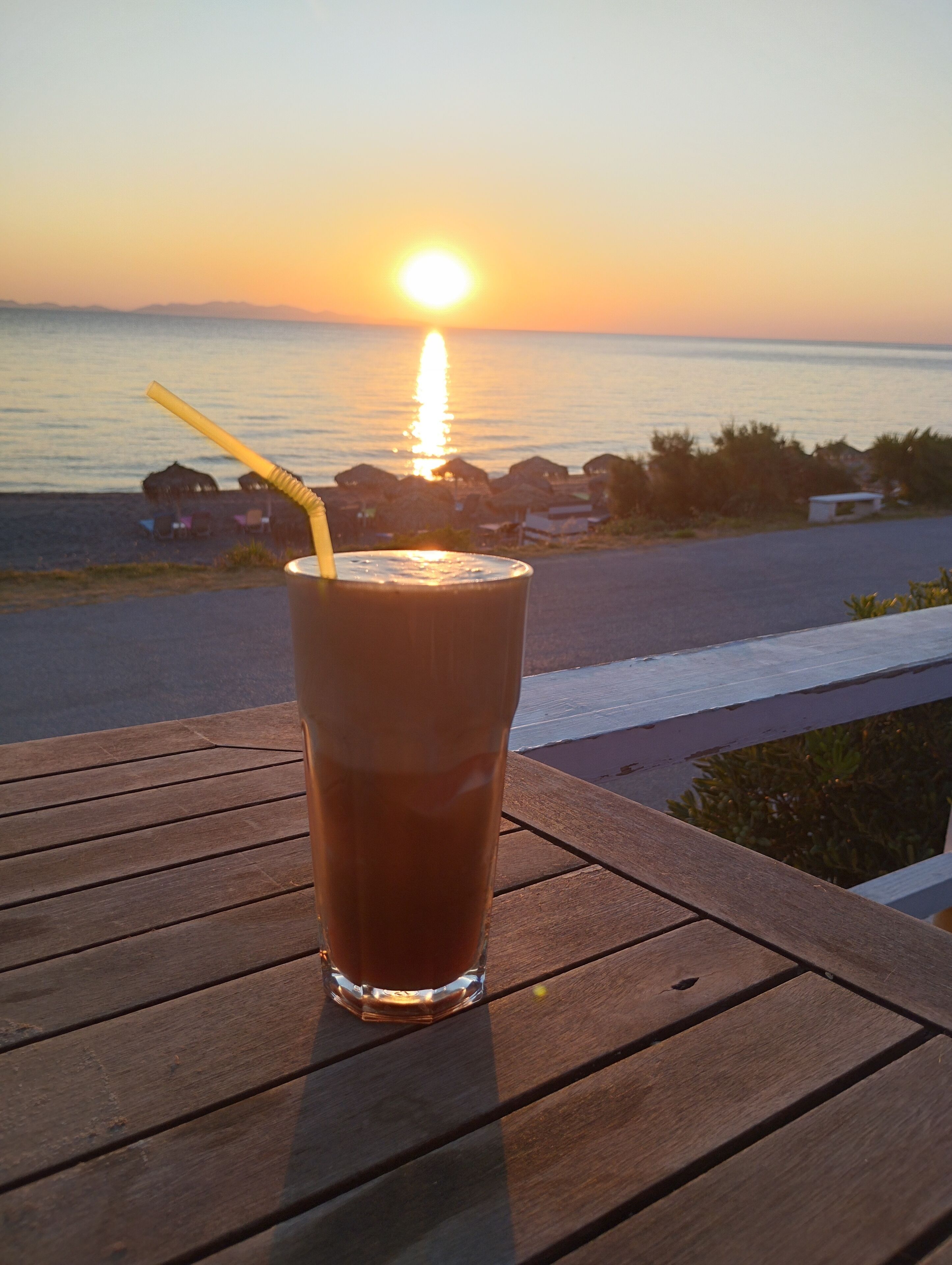 A glass of iced coffee on a wooden table with a sunset view in the background.