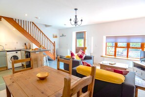 Eine moderne Küche mit integriertem Waschmaschinenanschluss, einem Essbereich mit Holztisch und Stühlen, einer Treppe sowie einem Wohnbereich mit Sofa und Kissen.