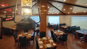 A restaurant with wooden tables and chairs, a hanging light fixture, and a view of the sea through large windows. - Carom The Castle Resort Fagu Shimla (Theog)