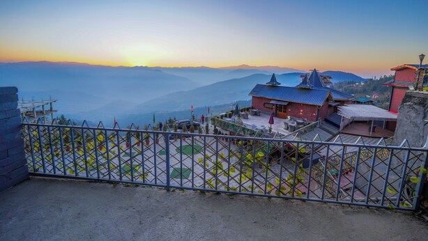 A building with a traditional roof, a fenced courtyard with potted plants, and a view of distant mountains. - Carom The Castle Resort Fagu Shimla (Theog)