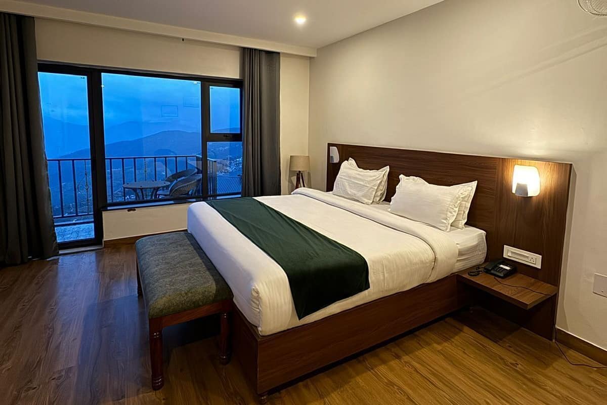 Een moderne hotelkamer met een groot bed, een bankje, een nachtkastje met een lamp en uitzicht op de stad door een groot raam.