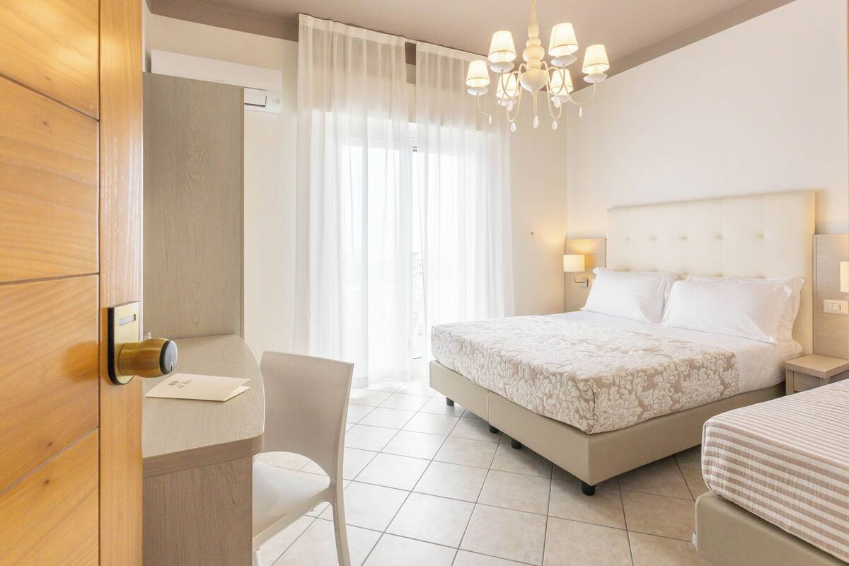 Habitación de hotel con cama, mesitas de noche, una silla, escritorio, candelabro y ventana con cortinas.