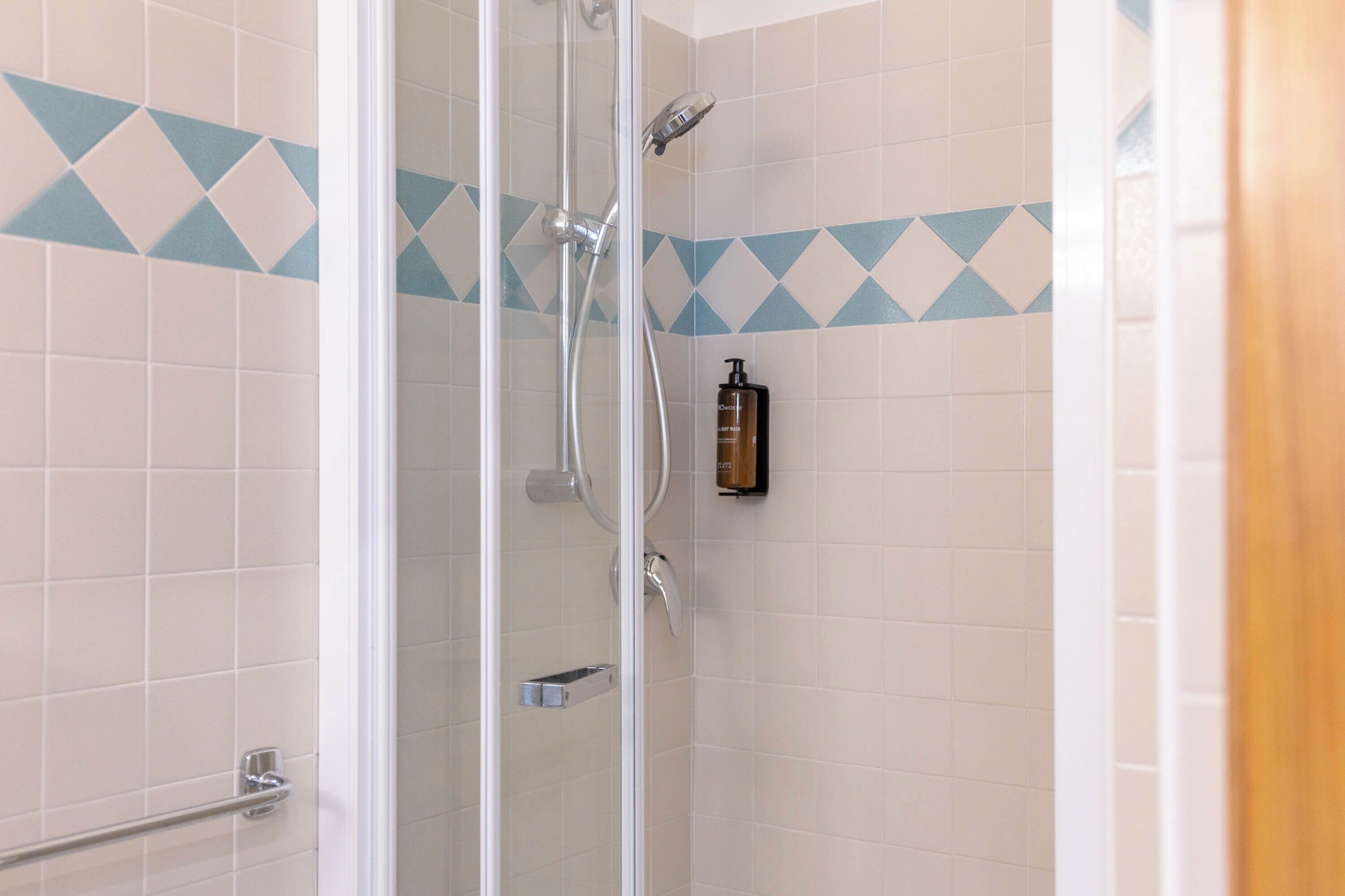 A shower with a glass door, a hand dryer, and a bottle of shampoo on the wall.