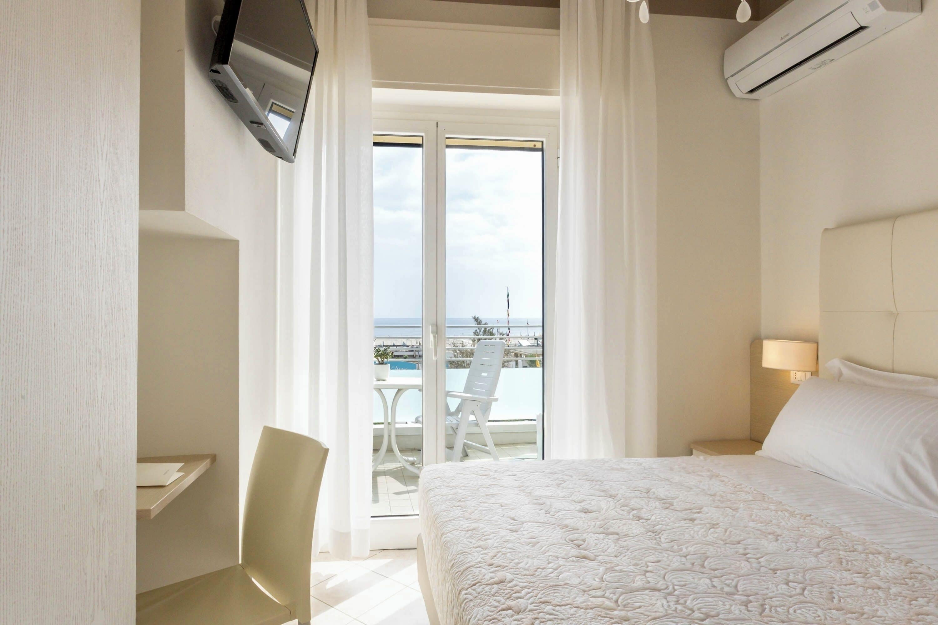 A hotel room with a bed, a desk, a chair, a TV, an air conditioner, a lamp, and a balcony with a view of the sea.