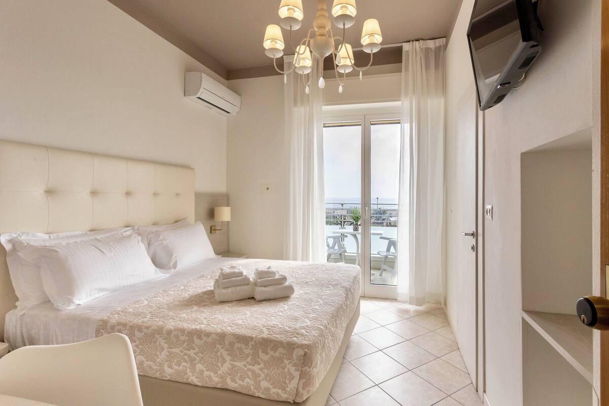 Habitación de hotel con cama, televisor, candelabro y balcón con vistas.