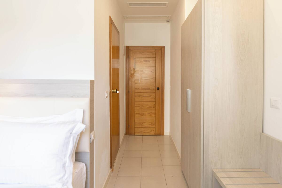 Un pasillo de hotel con una puerta de madera, una cama con ropa blanca y un banco.